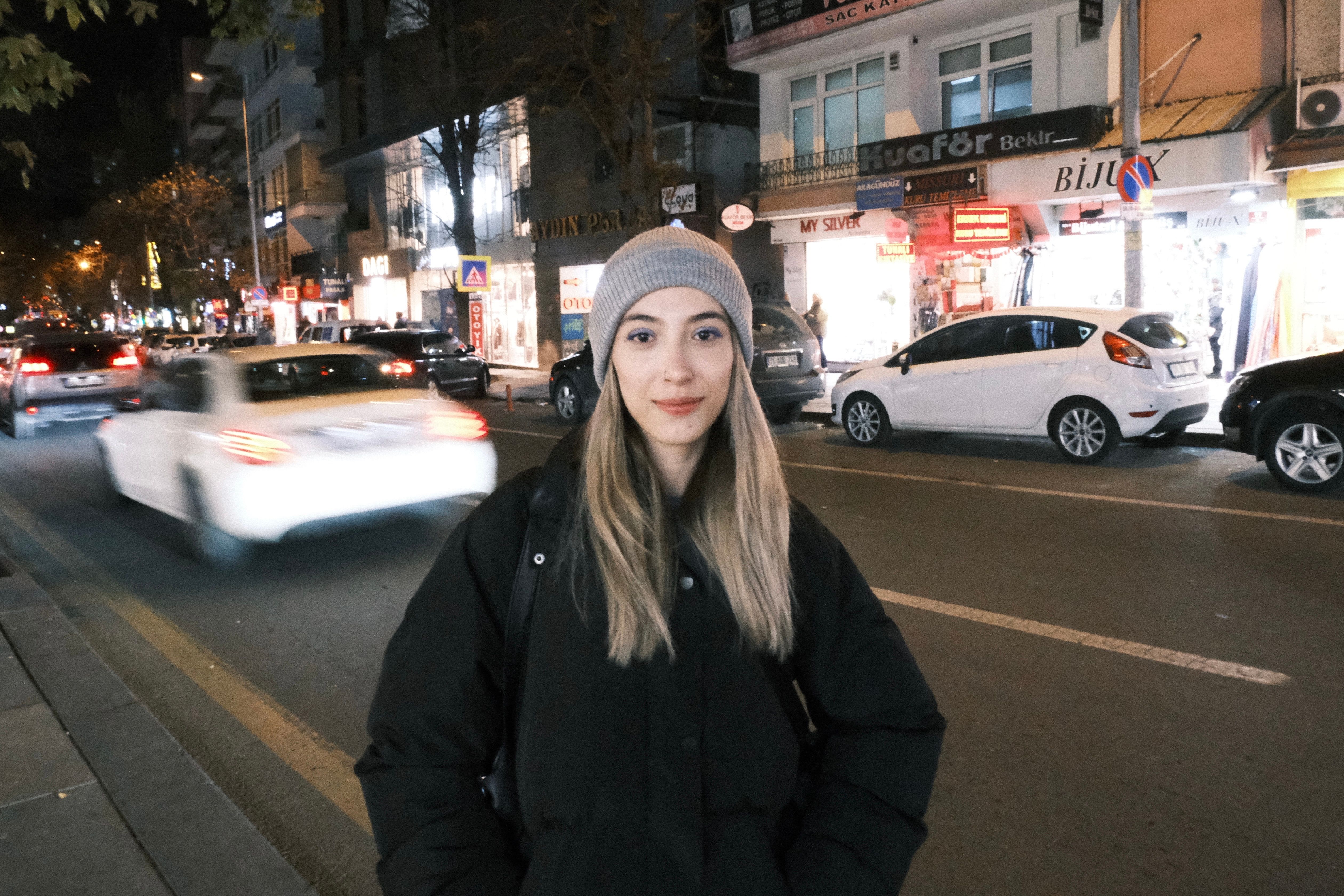 Blonde woman in a jacket and beanie on a bustling city street at night.