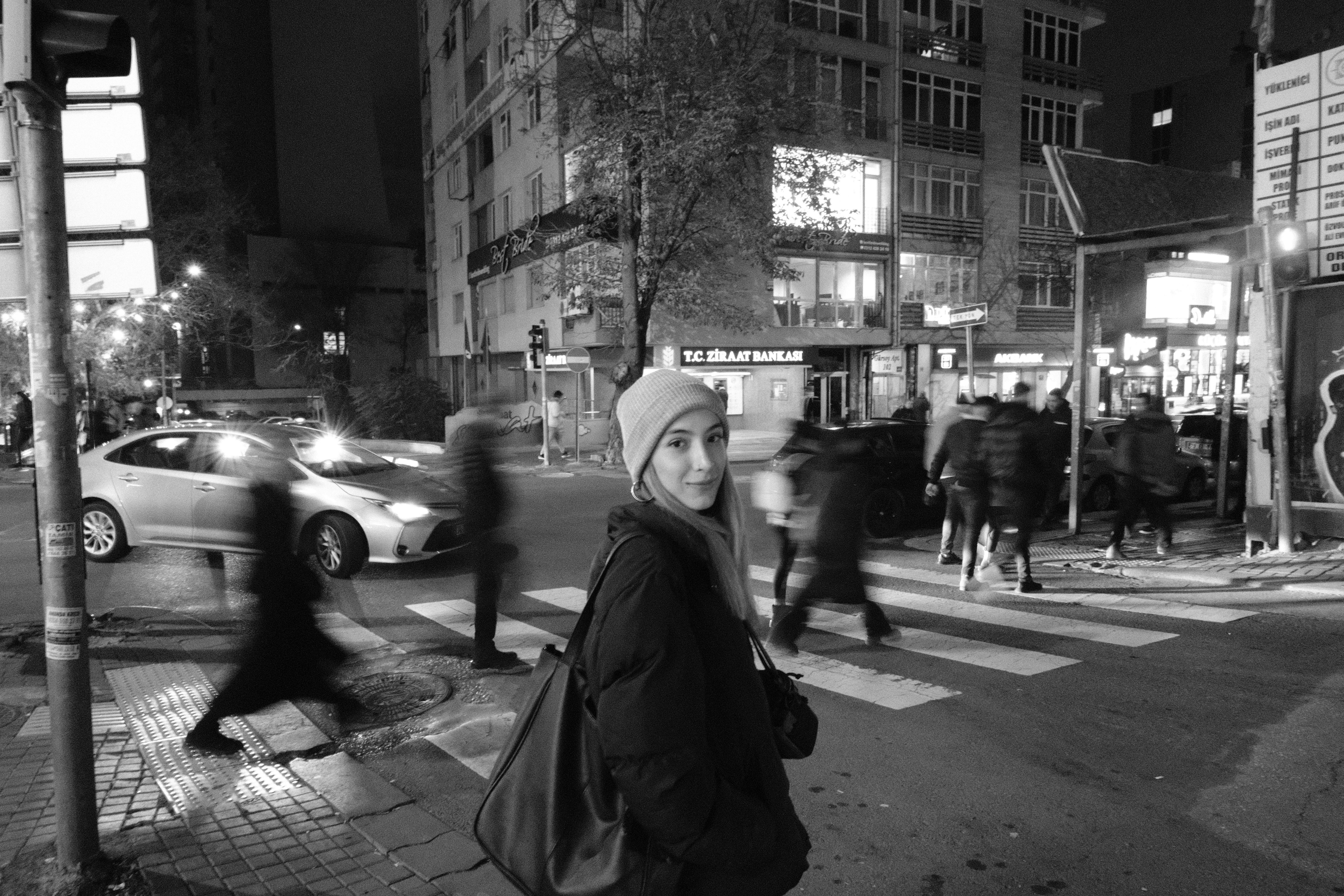 Black and white image of a woman at a crosswalk in a city street at night.