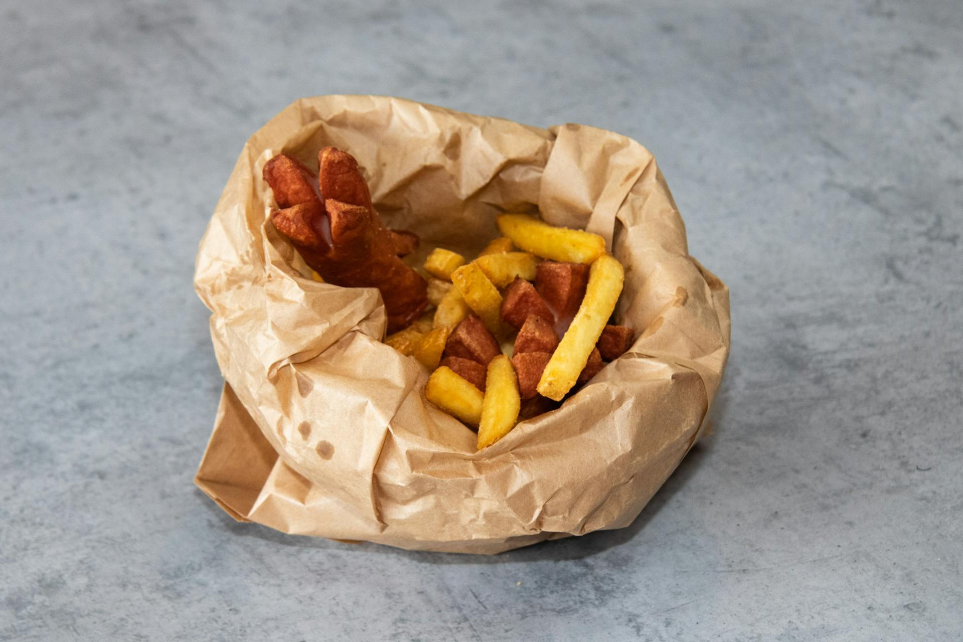 A delicious serving of crispy fries and sausages in a kraft paper bag on a gray background.