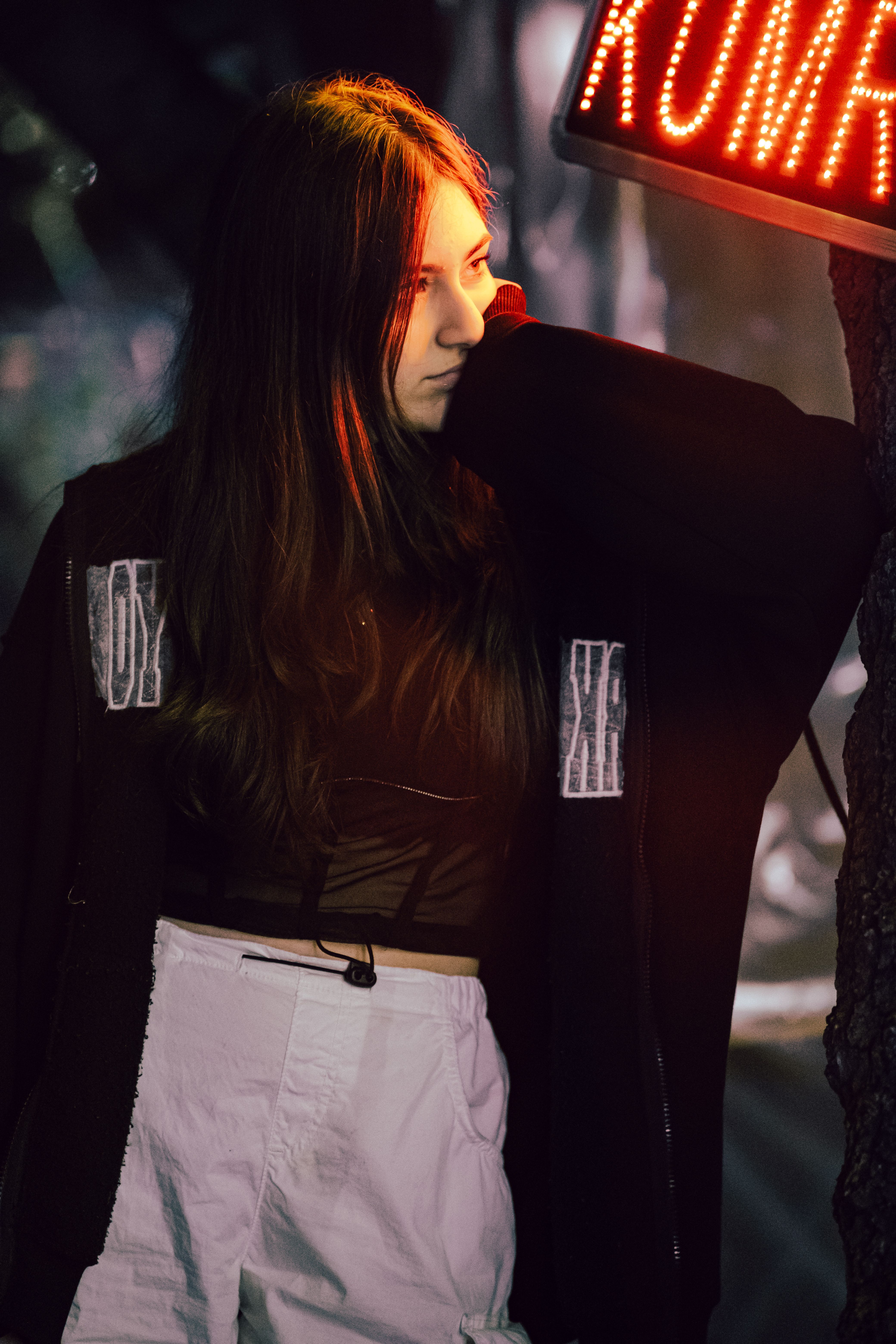 A young woman in street clothes stands pensively under neon lights.