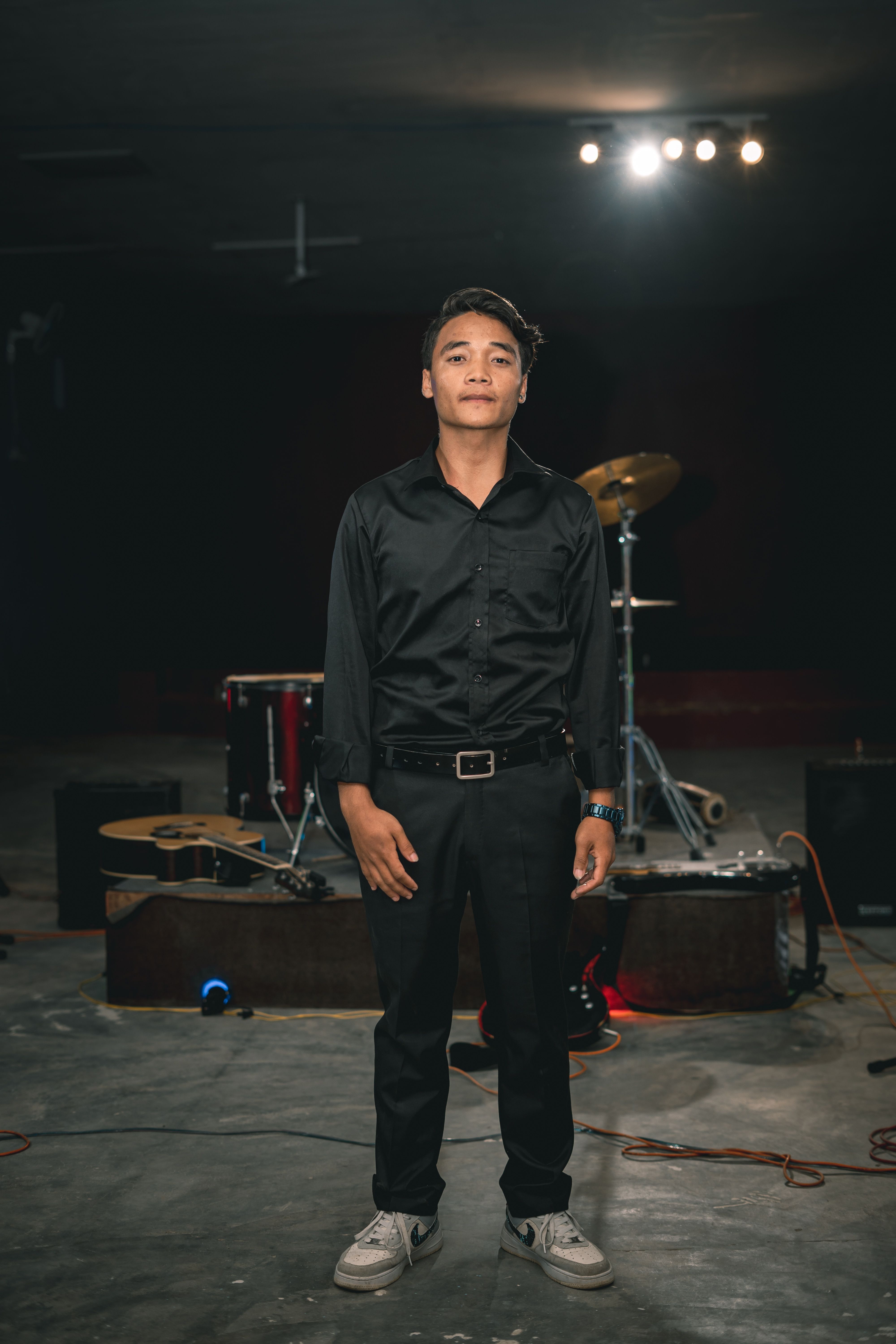 Adult male musician standing on stage with drums and guitar in dim light.