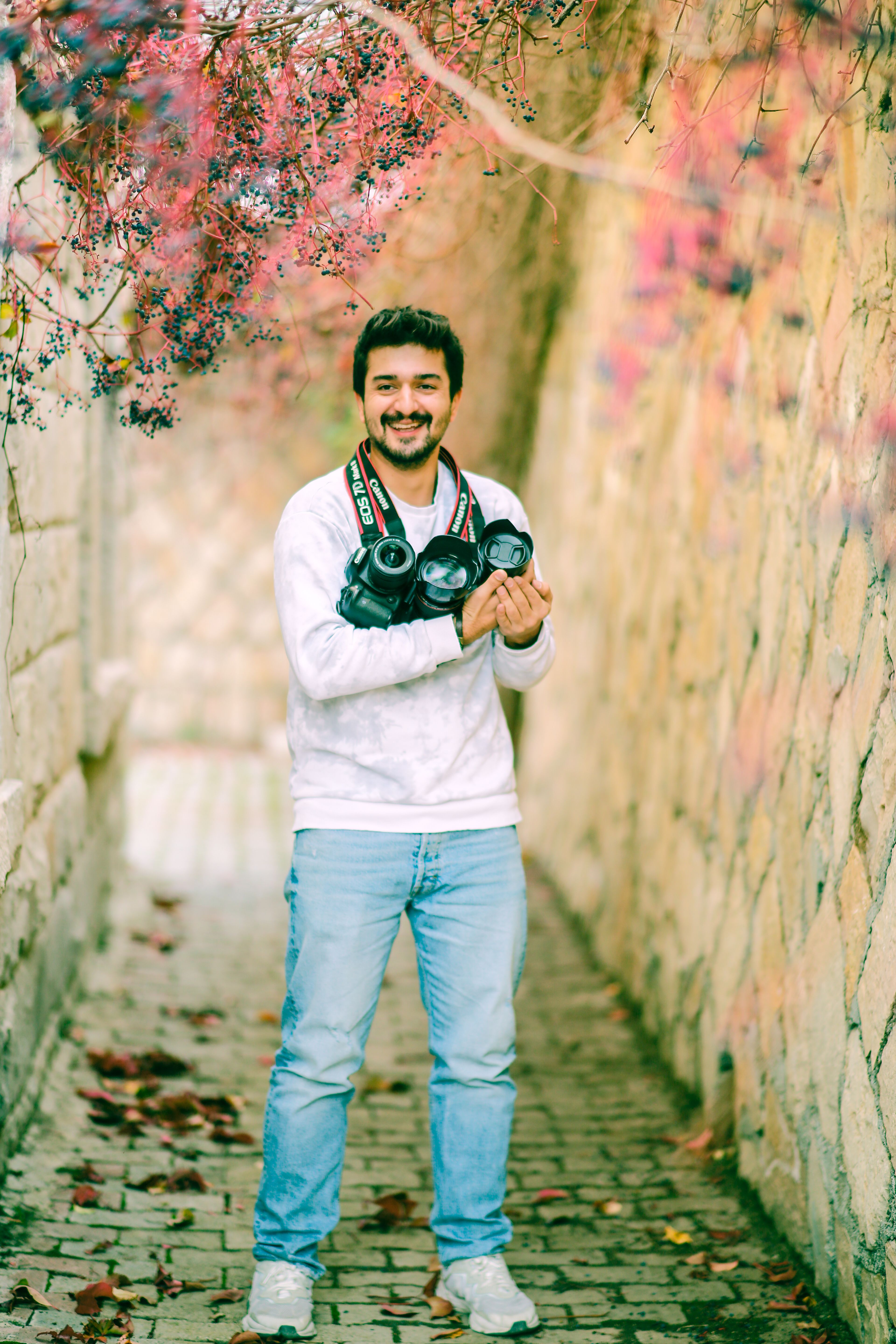 A smiling man with short hair stands in a scenic alley holding multiple cameras.