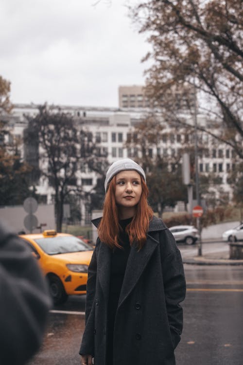 Young Woman in a Coat and Hat Standing on the Street in City 