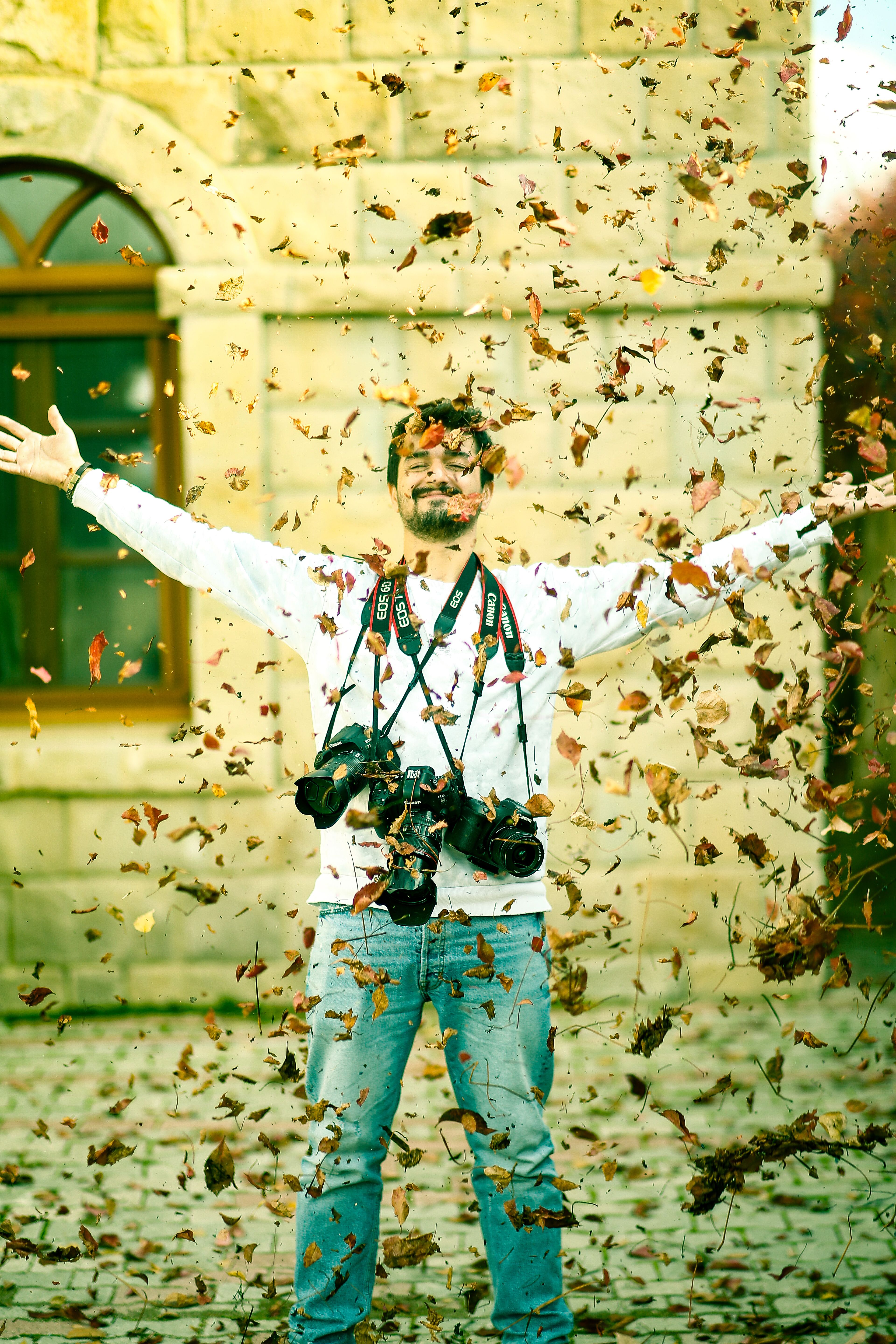 A man joyfully poses with arms raised among falling autumn leaves.