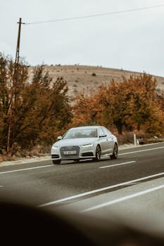 A silver sedan travels on a rural road surrounded by autumn foliage, depicting a serene journey.