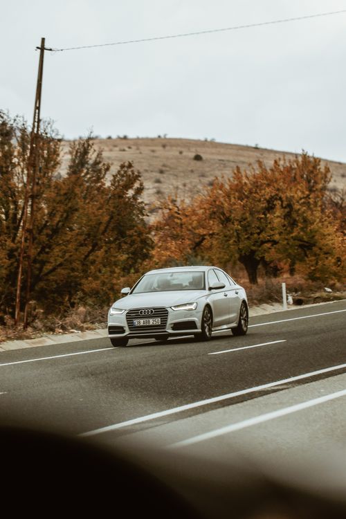 Free A silver sedan travels on a rural road surrounded by autumn foliage, depicting a serene journey. Stock Photo
