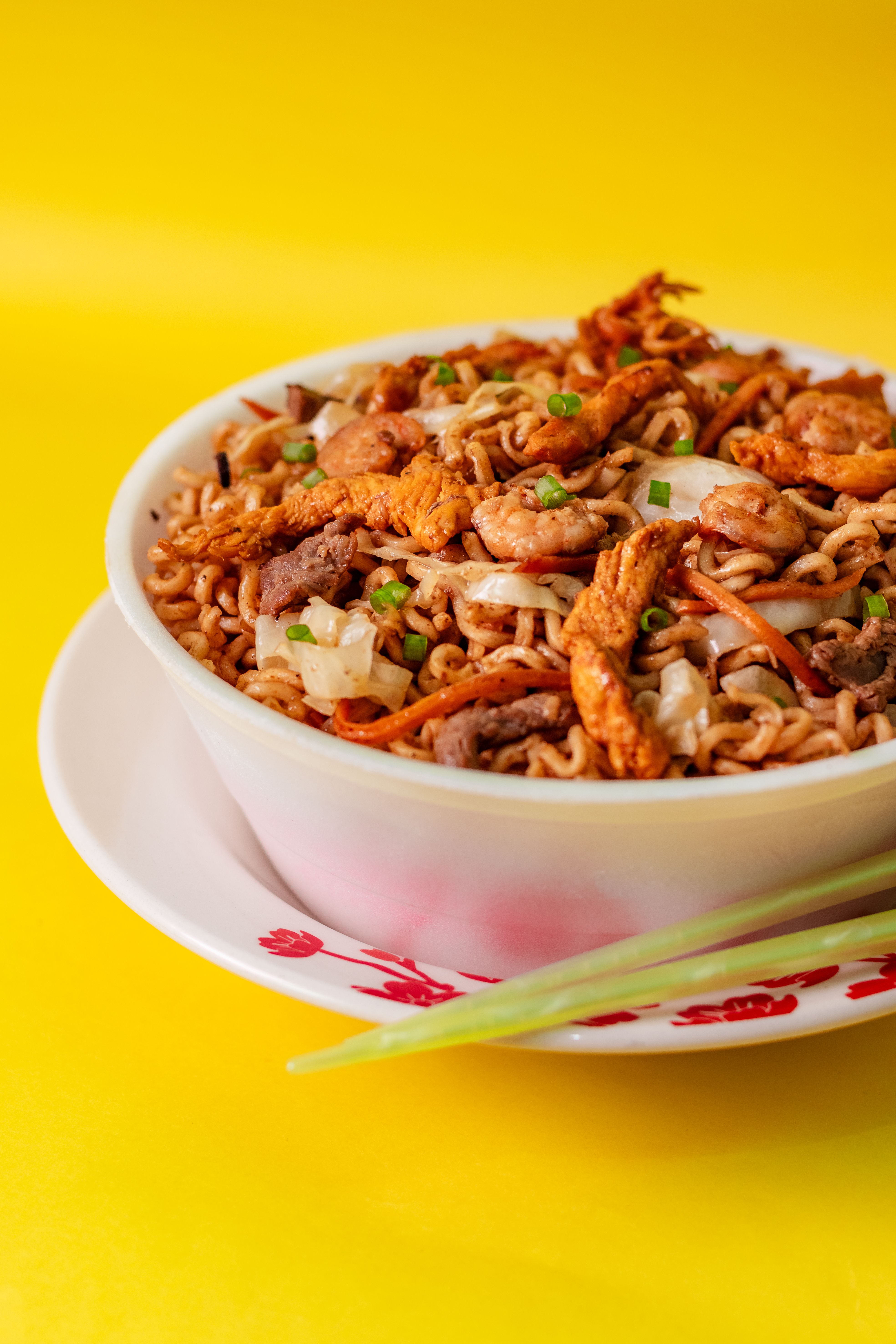 Savor a bowl of stir-fried noodles with tender chicken and fresh vegetables on a vibrant yellow backdrop.
