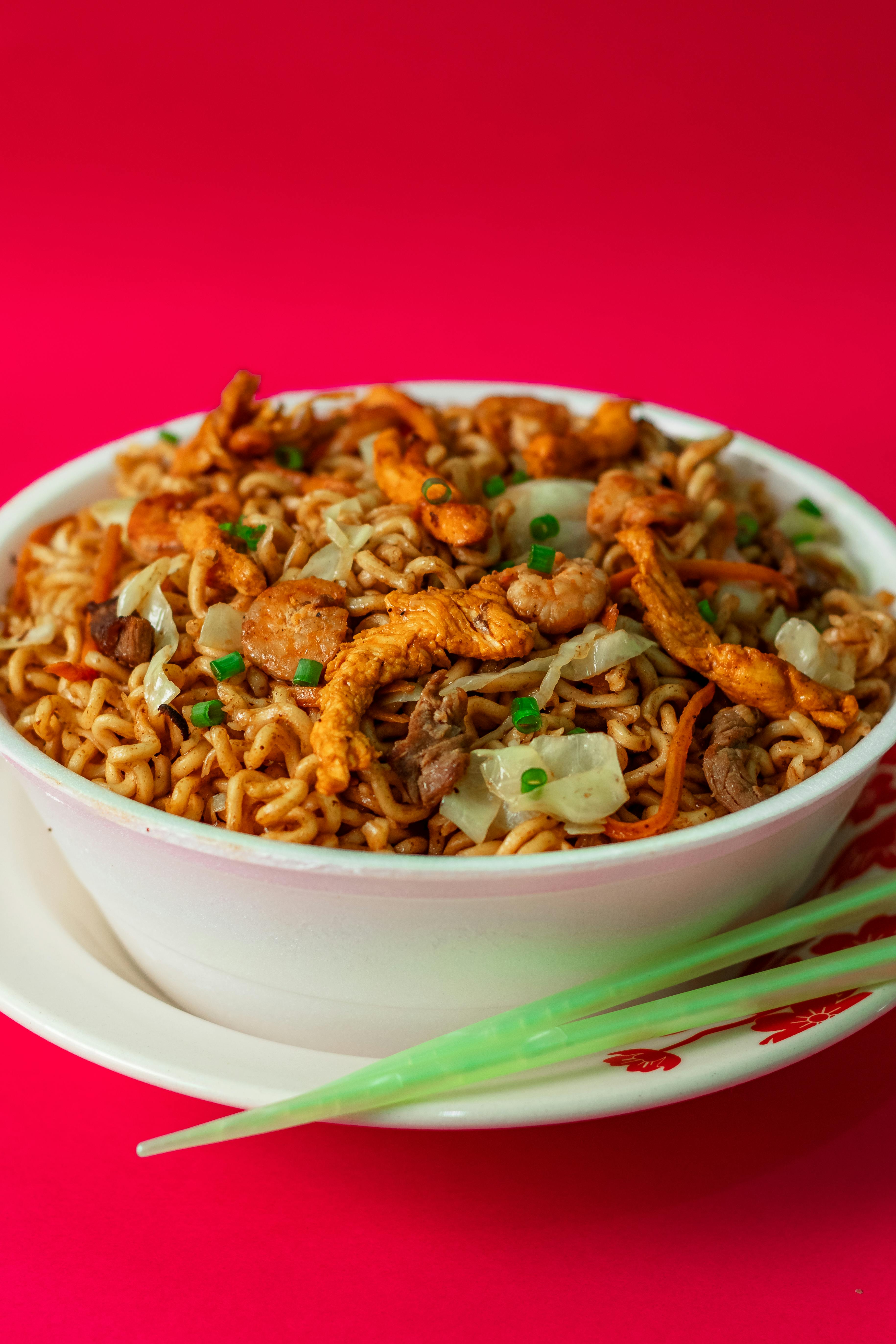 A bowl of golden stir-fried instant noodles mixed with red tomatoes, scrambled eggs, and green scallions, showing the colorful Nigerian-style preparation