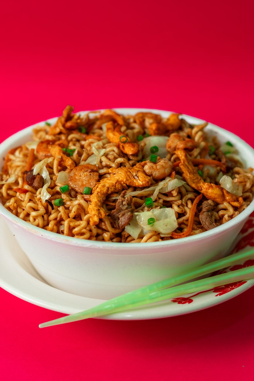 A bowl of golden stir-fried instant noodles mixed with red tomatoes, scrambled eggs, and green scallions, showing the colorful Nigerian-style preparation