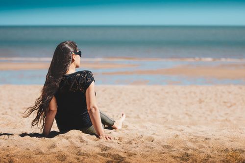 Free Woman with long hair sitting on a sunny beach, enjoying the ocean view. Stock Photo