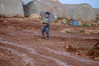A young child walks through a muddy refugee camp in Idlib, Syria, amidst tents and rustic surroundings.