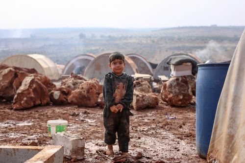 Free A young boy stands in a muddy refugee camp in Idlib, Syria, amidst makeshift tents and rocky terrain. Stock Photo