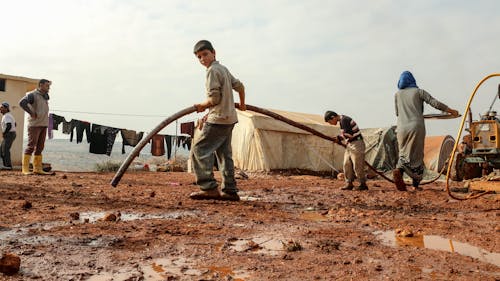 Boys Holding a Hose on a Muddy Ground at a Refugee Campsite 