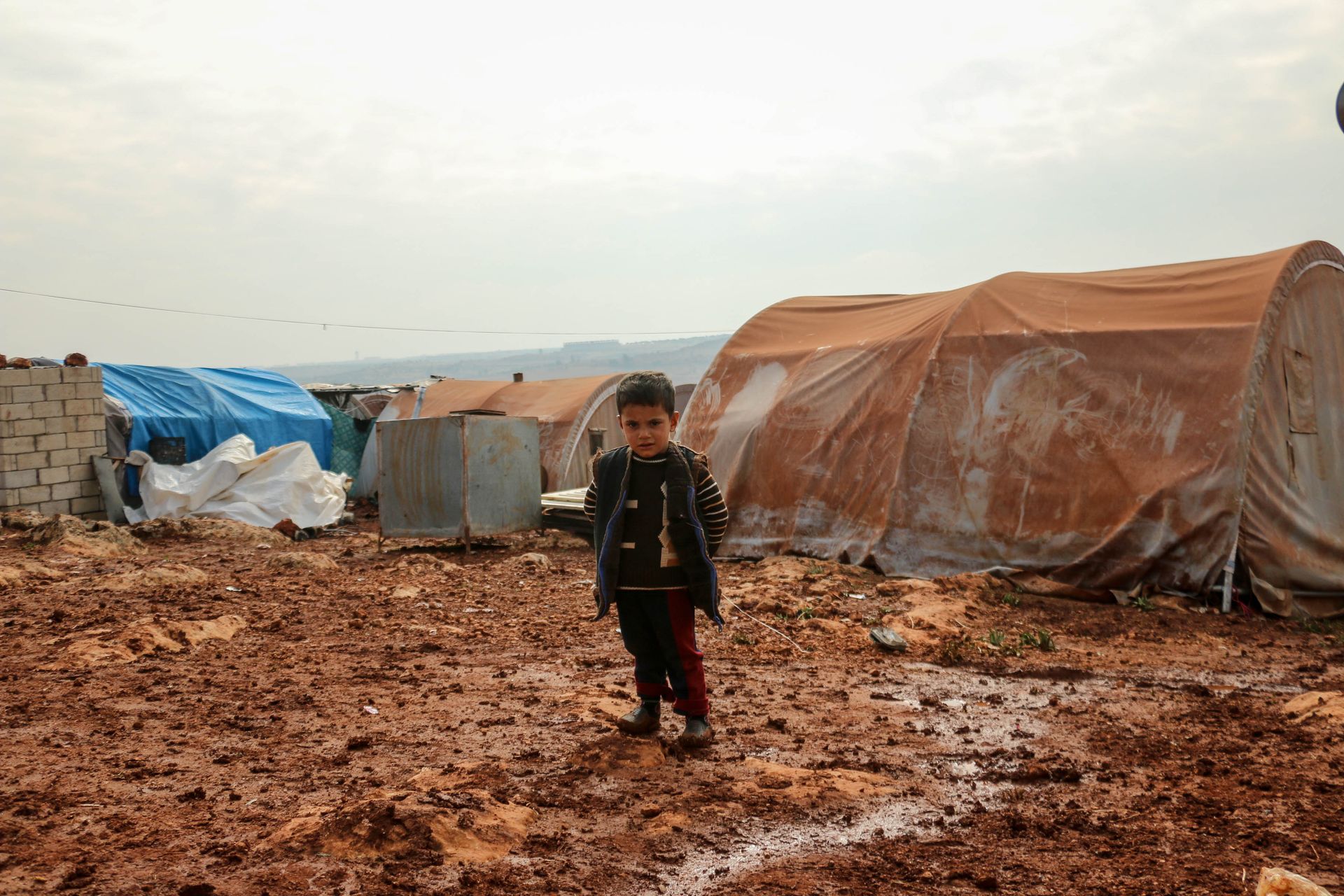 A young boy stands in a Syrian refugee camp, surrounded by tents under a cloudy sky.