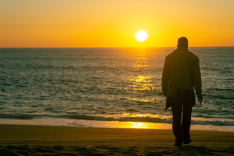 Man Walking On The Beach Towards The Sea At Sunset