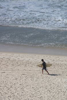 Aerial view of a man walking with a surfboard on a sandy beach in Rio de Janeiro, Brazil.