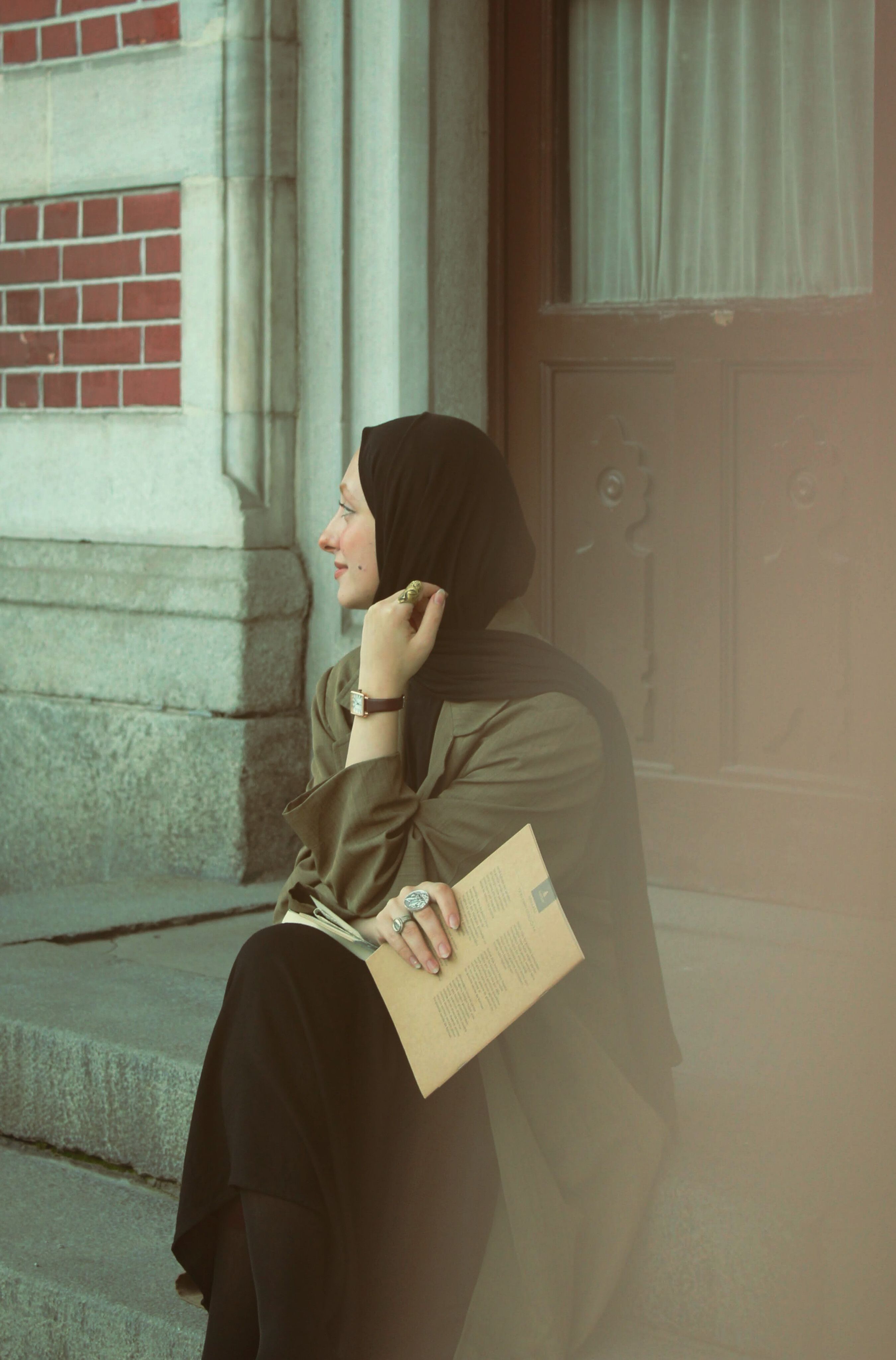 Young woman in hijab sits on steps, holding a book and looking away thoughtfully.