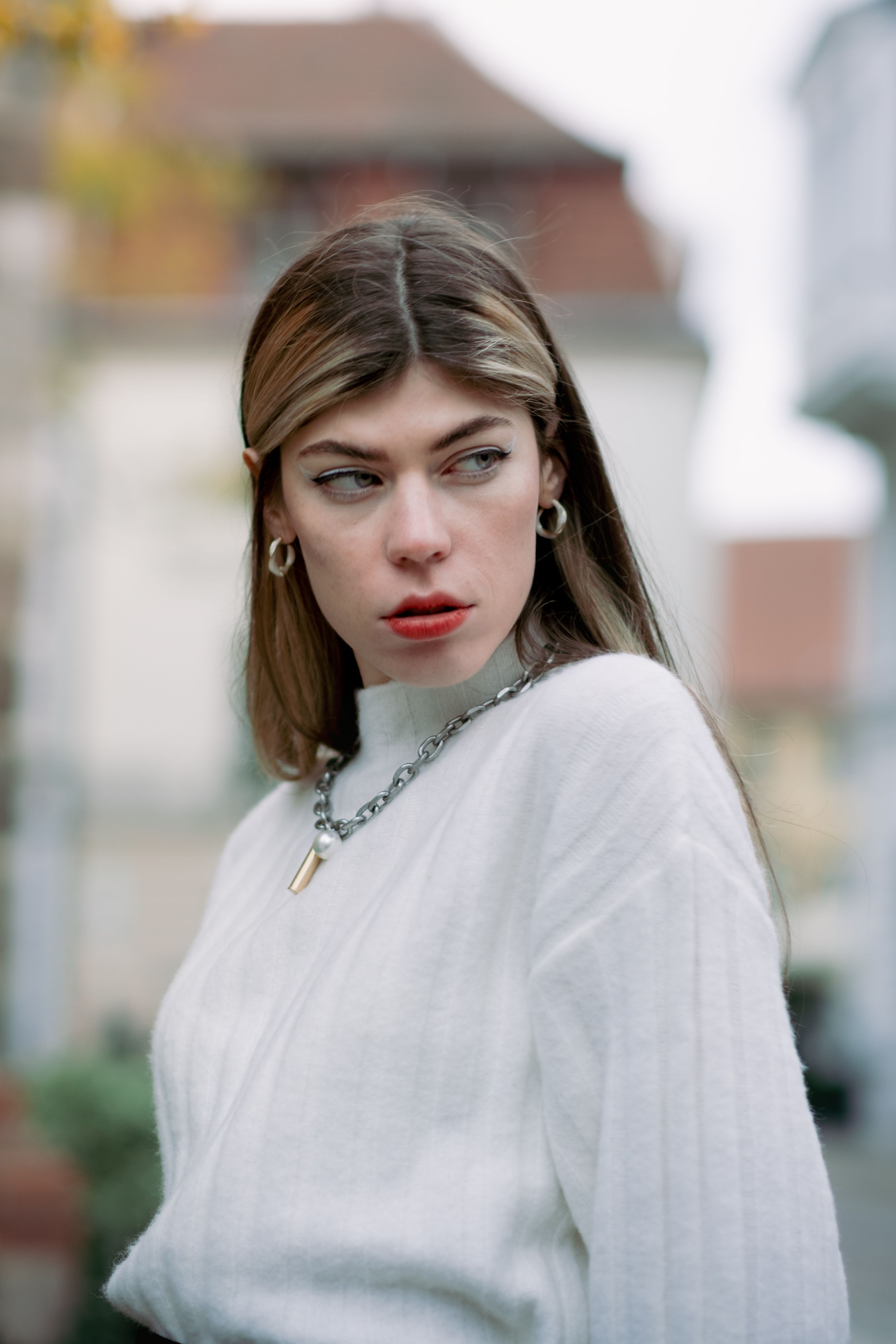Elegant young woman with blonde hair posing outdoors wearing a white sweater and necklace.