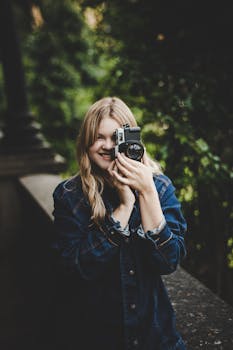 Smiling woman in denim jacket taking a photo outdoors with SLR camera.