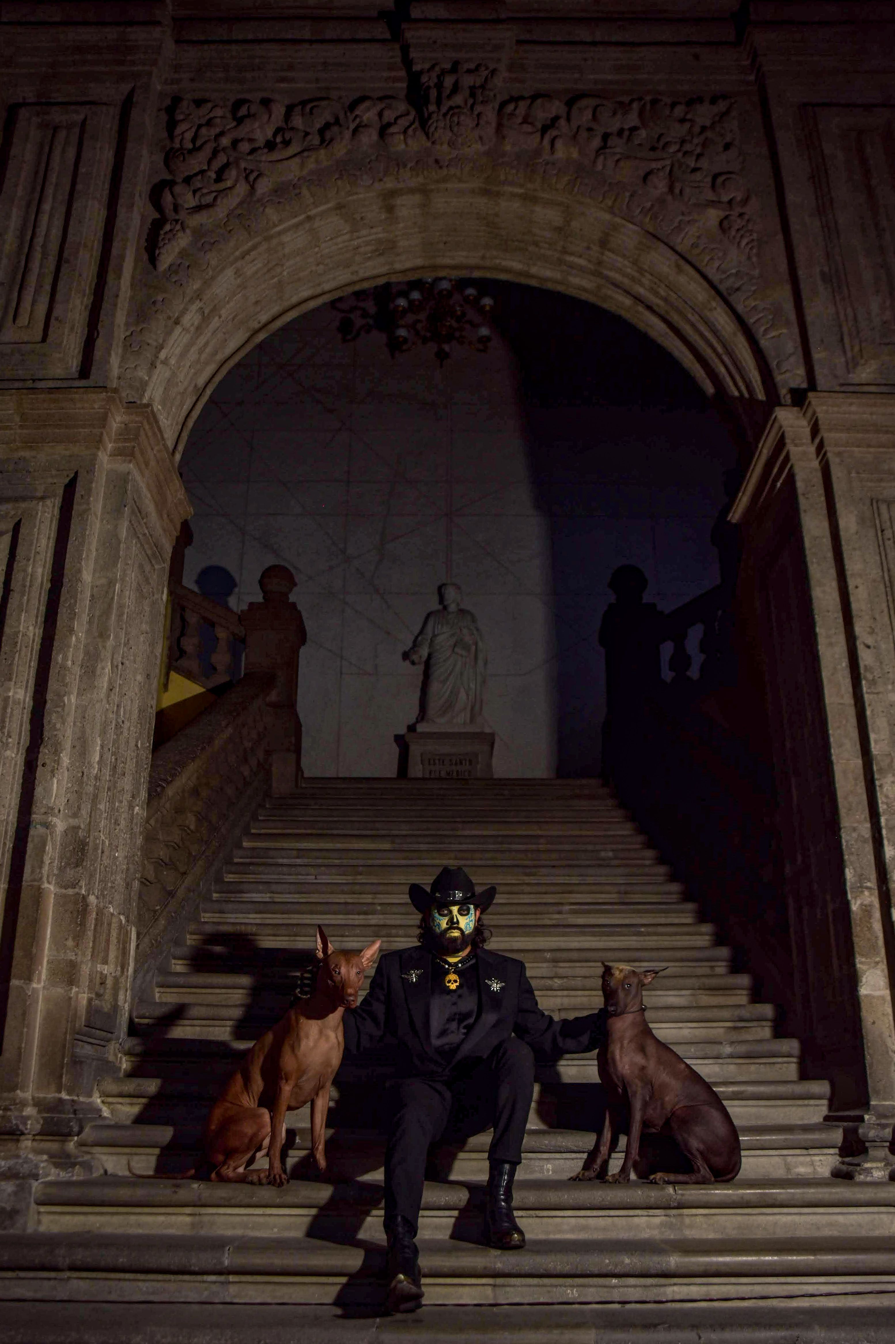 A man in traditional Día de Muertos costume sits on stairs with two dogs in an urban setting at night.