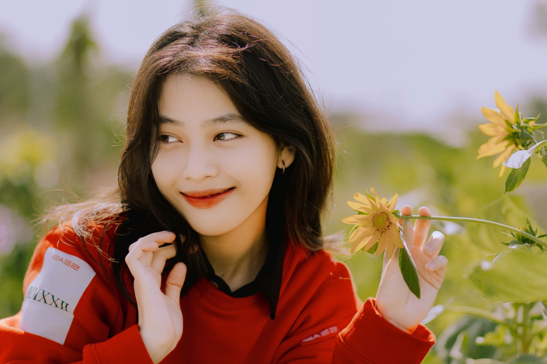 A young woman in a red sweater smiles, holding a sunflower in a sunny outdoor field.