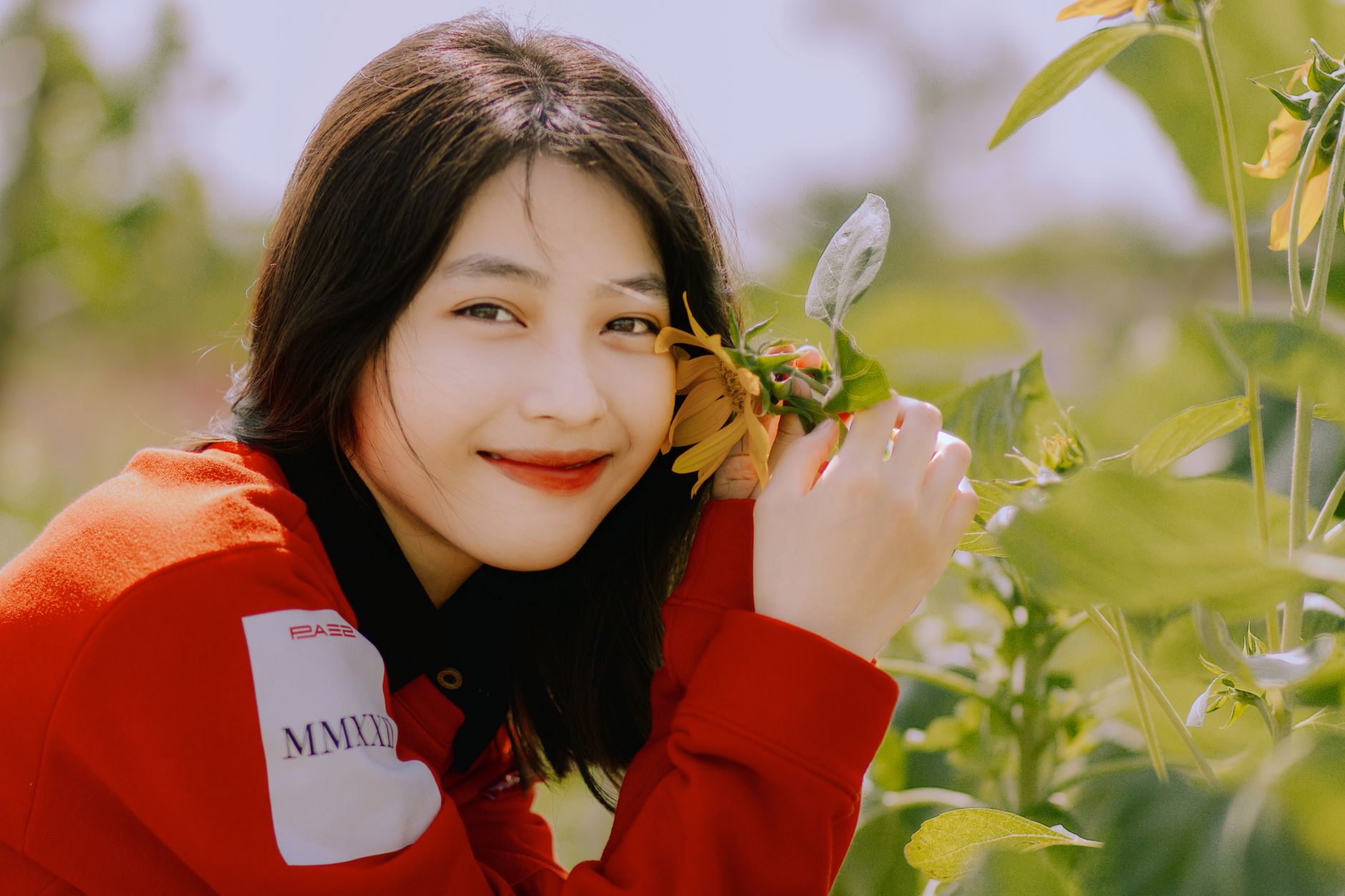 Asian woman with long black hair smiling and holding a sunflower in a garden setting.