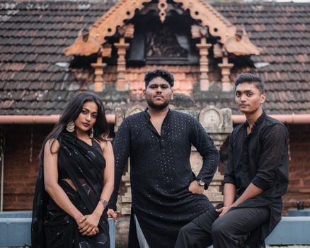 Three adults in traditional Indian attire pose elegantly in front of a cultural building.