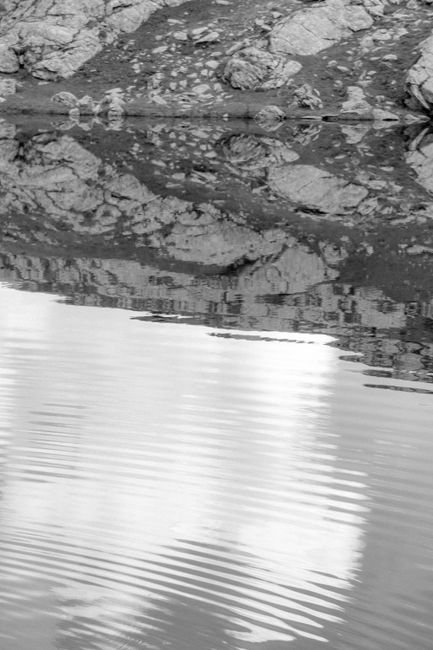 Black and white image of a tranquil lake with rocky reflections and ripples.