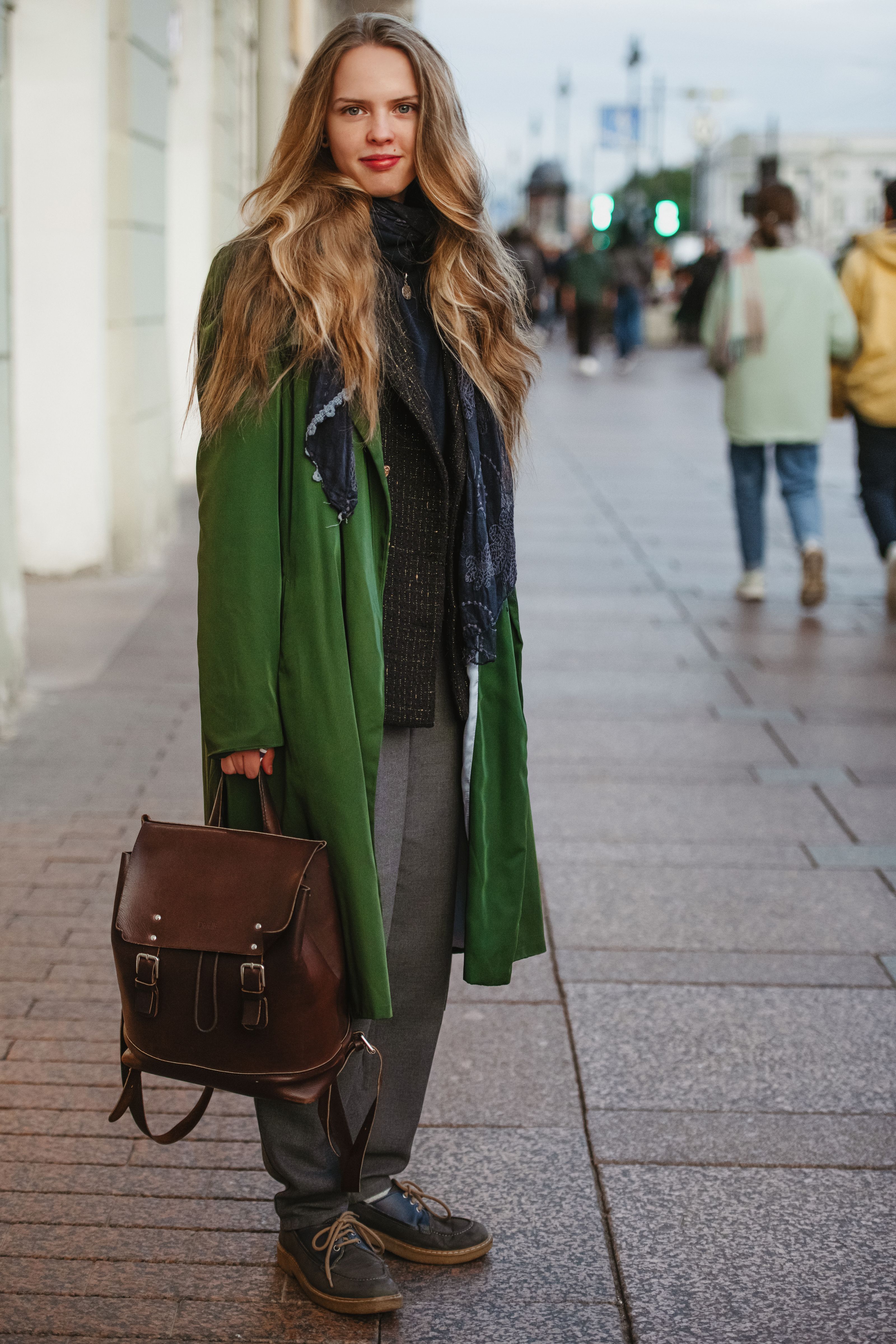 Mulher fashionista com longos cabelos loiros, usando casaco verde e segurando uma mochila de couro em uma rua urbana.