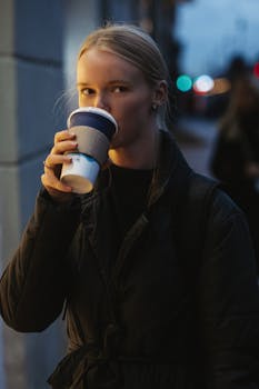 Portrait of a young woman drinking coffee on a city street at dusk.