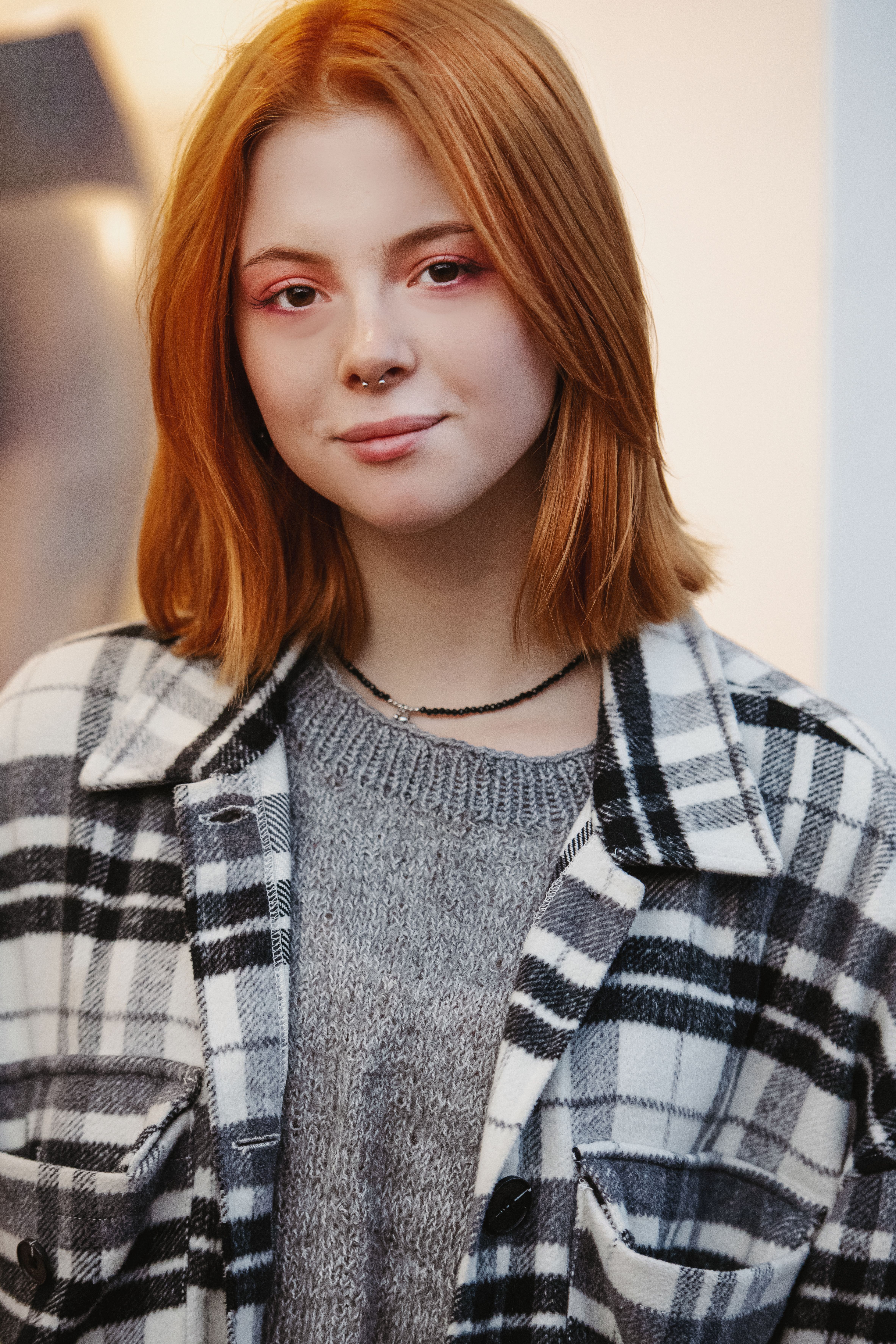 Portrait of a young redhead woman wearing a plaid jacket and gray sweater indoors.