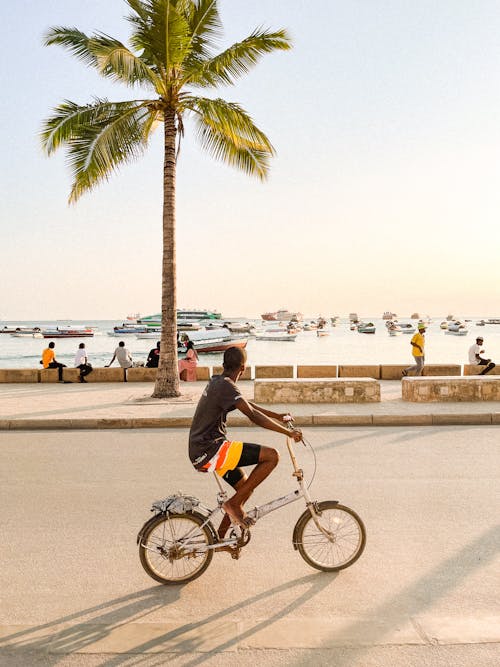 Young Man Riding a Bicycle Along the Coast Looking at the Boats Anchored by the Shore