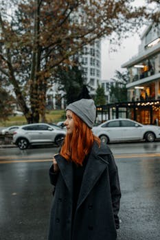 Stylish redhead woman with gray coat and beanie walking on a rainy city street.