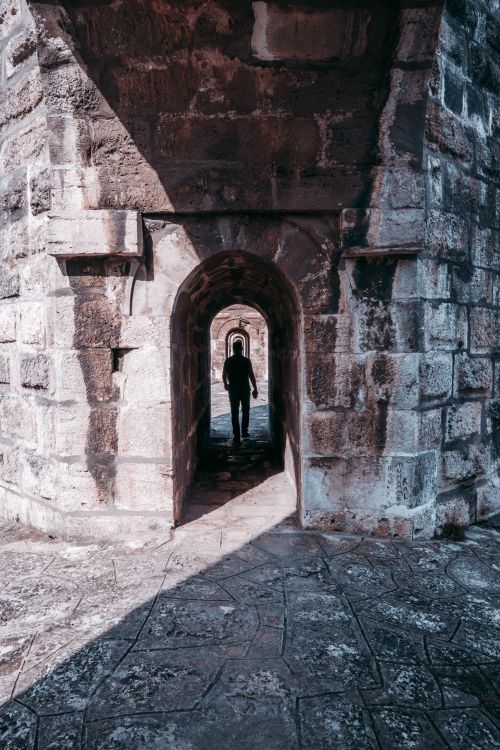 Free A silhouette of a person walking through a narrow historical stone tunnel with textured walls. Stock Photo