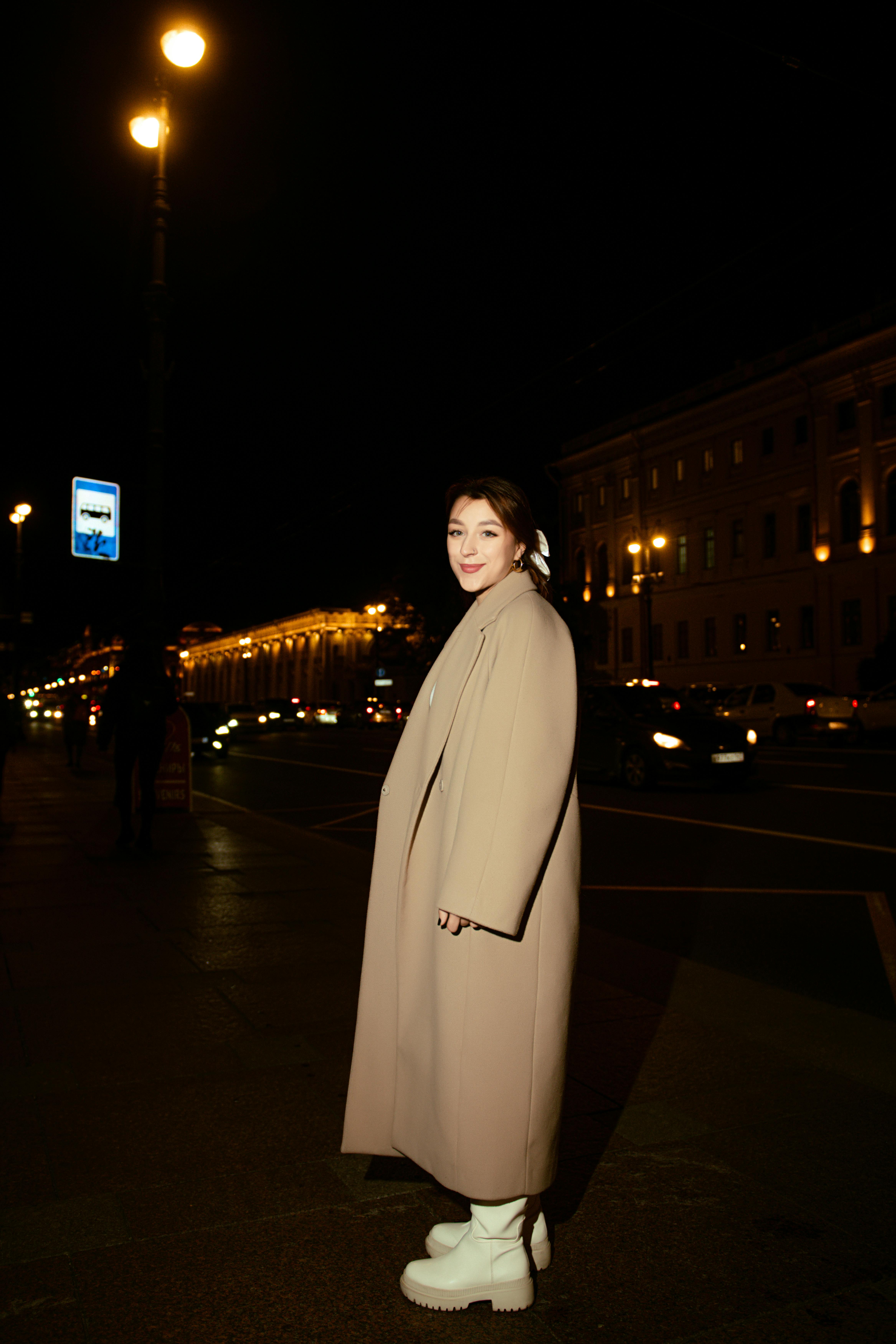 Stylish woman in beige coat and white boots poses on busy city street at night.