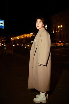Stylish woman poses in a beige coat on a vibrant city street at night, embracing urban fashion.