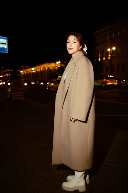 Stylish woman poses in a beige coat on a vibrant city street at night, embracing urban fashion.