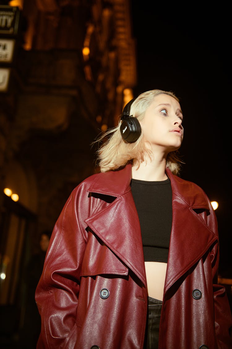 Young Woman In A Red Leather Coat Wearing Headphones And Walking Outside In The Dark 