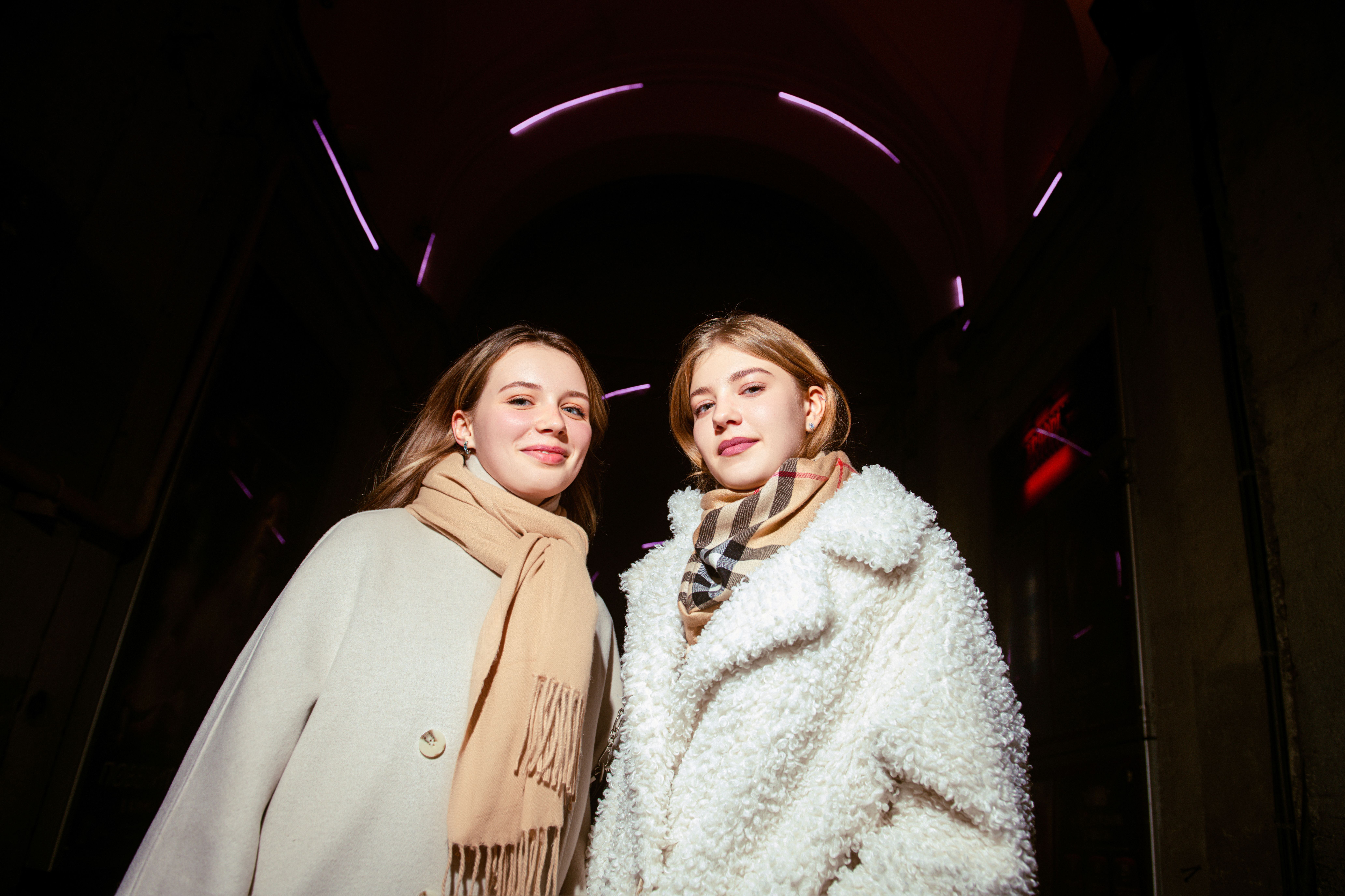 Two young women in cozy winter attire posing stylishly under neon lights.