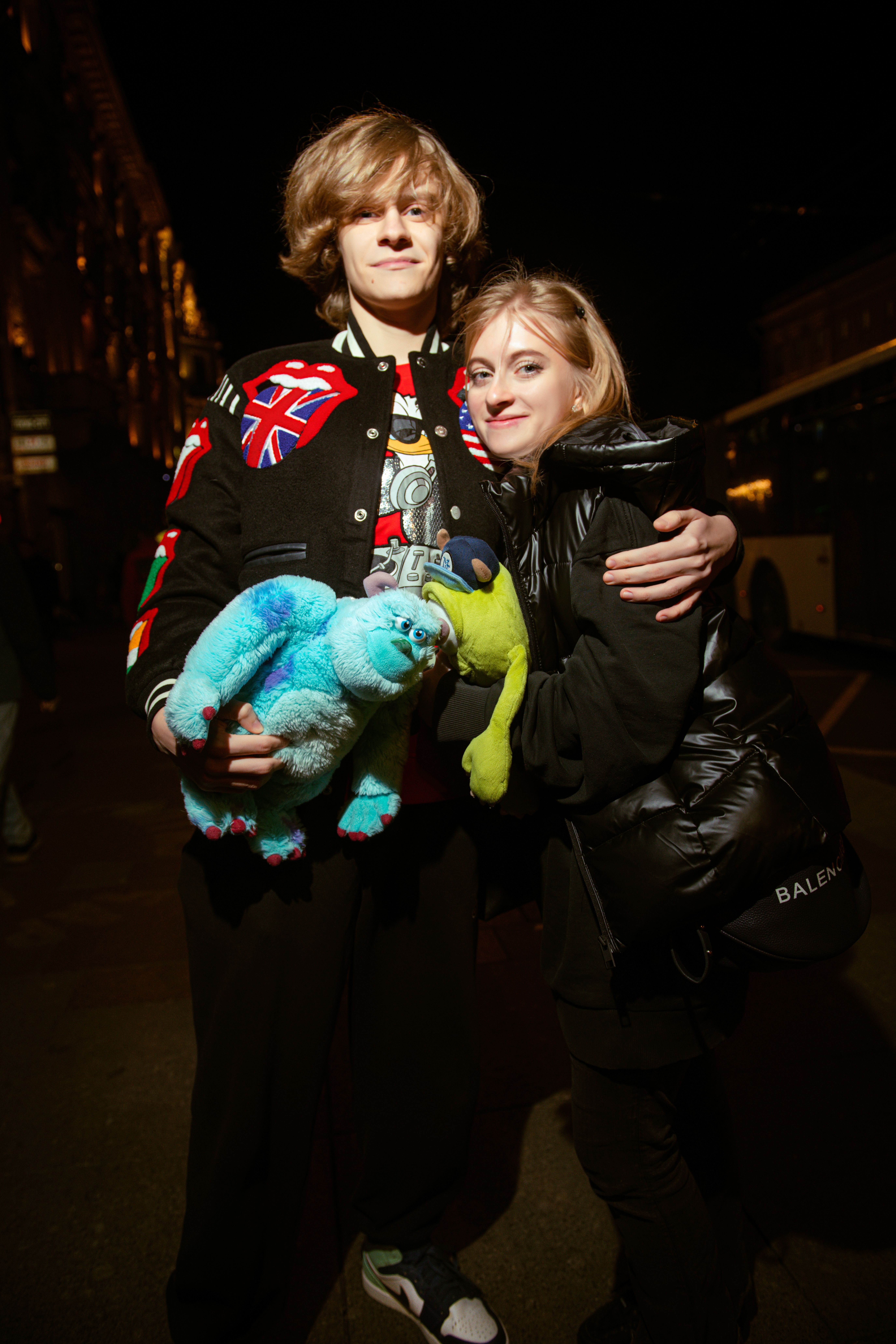 Happy young couple smiling and holding stuffed toys on a city street at night.