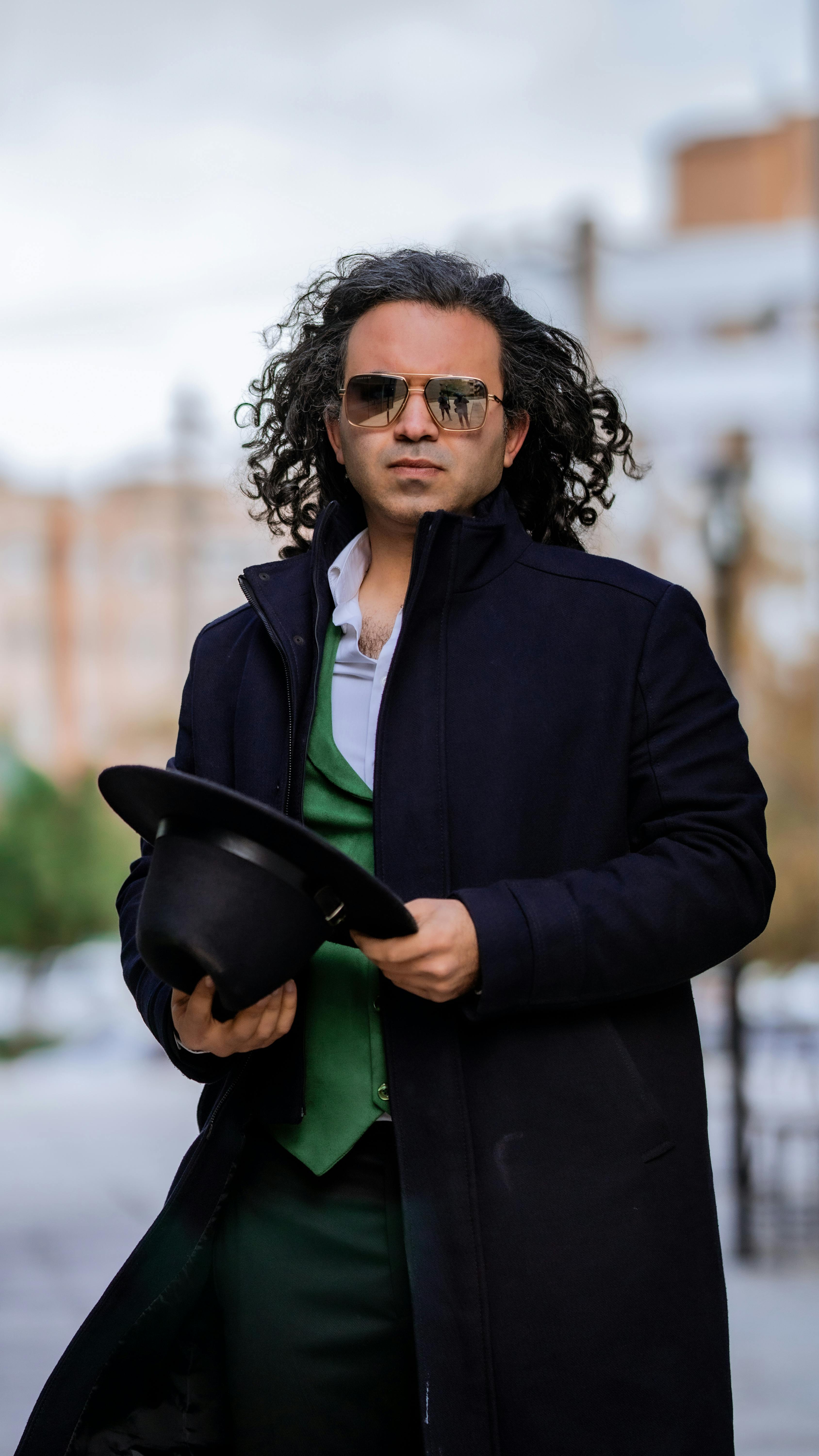 Elegant man with long hair and sunglasses in a black coat holding a hat, showcasing fashion style.