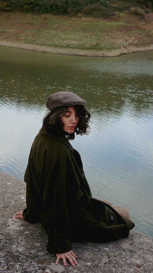 Woman in Flat Cap and Woolen Coat Sitting on Rock by River