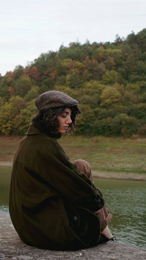 Woman in Flat Cap and Woolen Coat Sitting by River