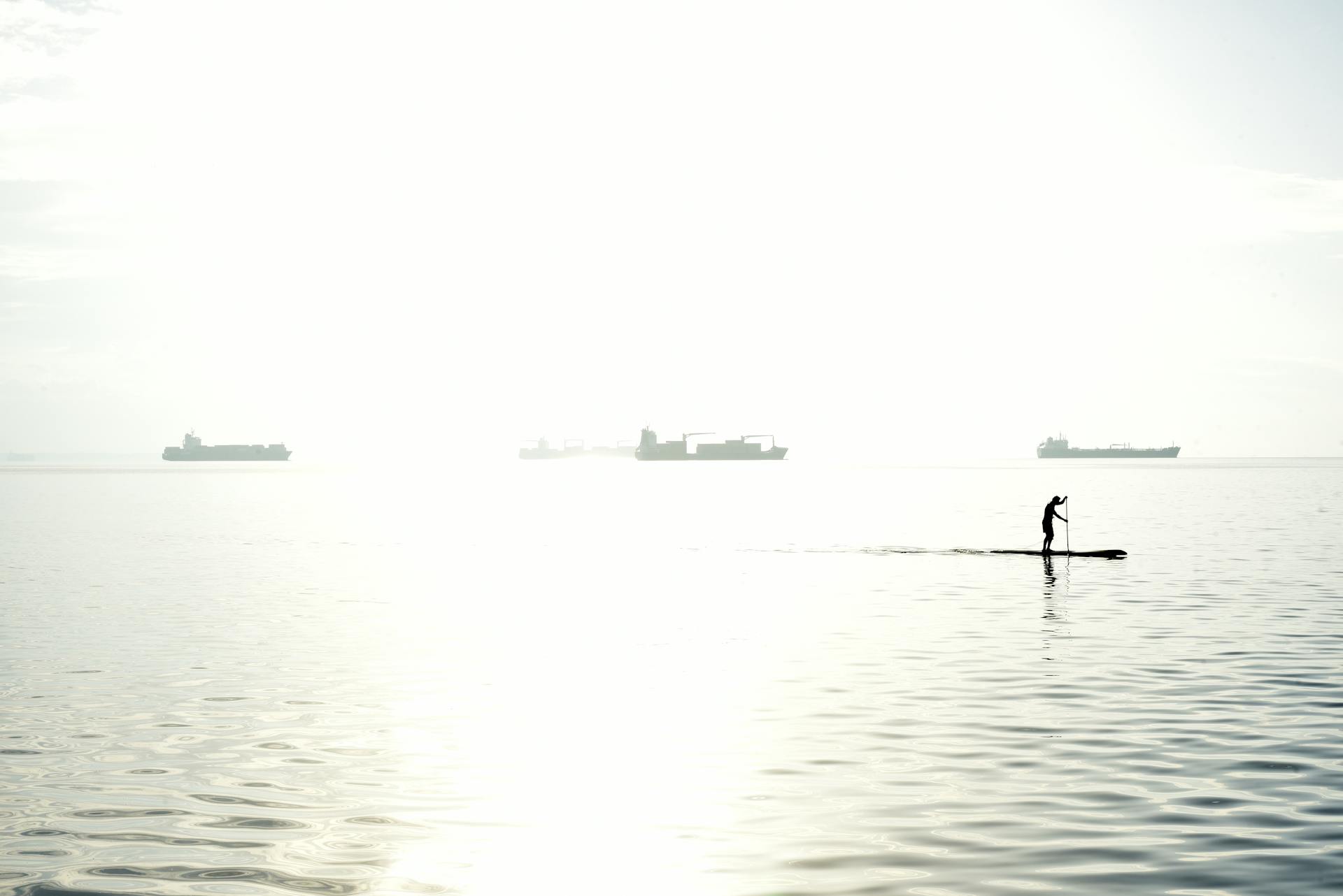 Man paddleboarding at sunset on peaceful Thessaloniki sea with ships in the distance.