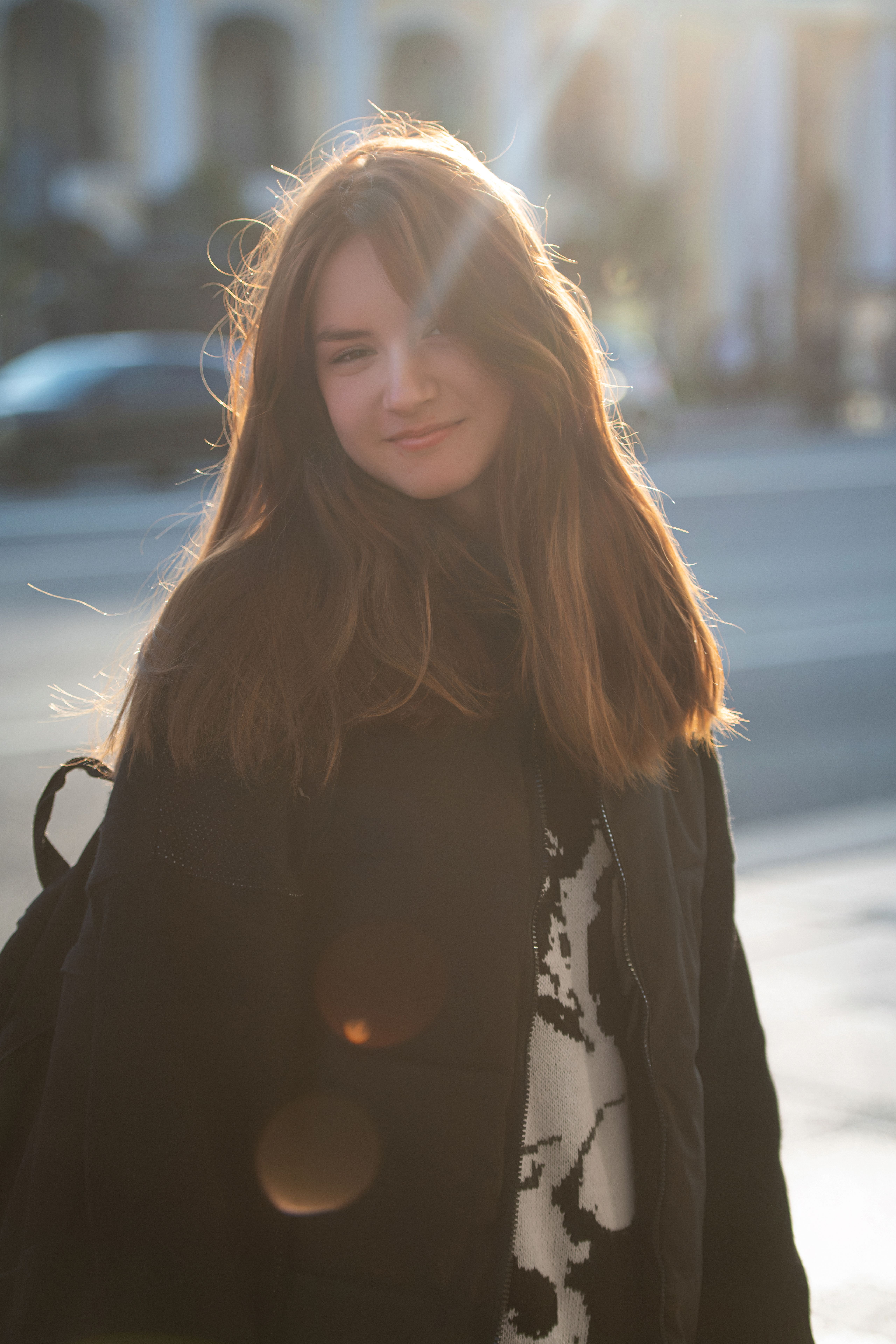 Young woman in a sunlit urban setting, wearing a black jacket and smiling.