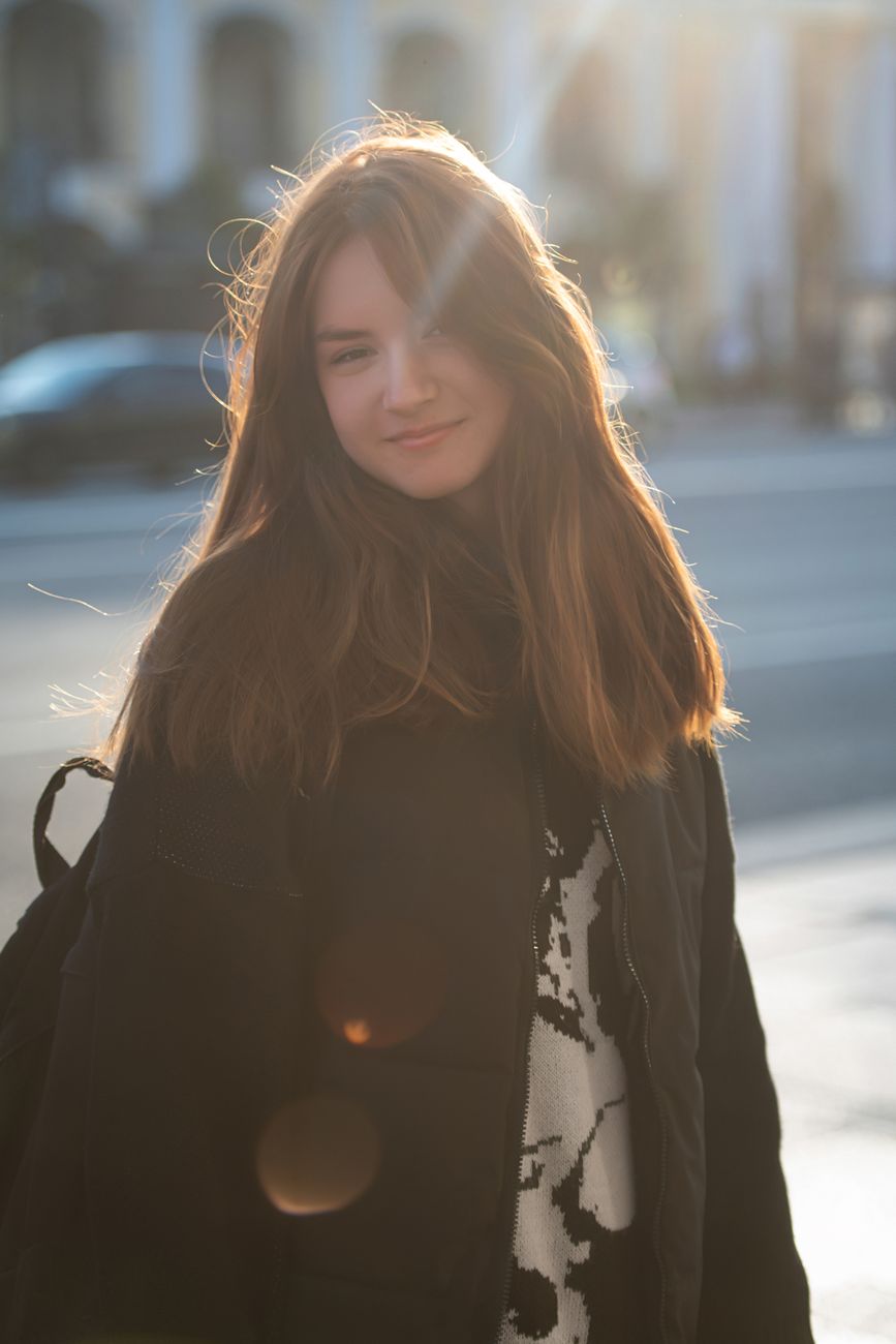 Young woman in a sunlit urban setting, wearing a black jacket and smiling.
