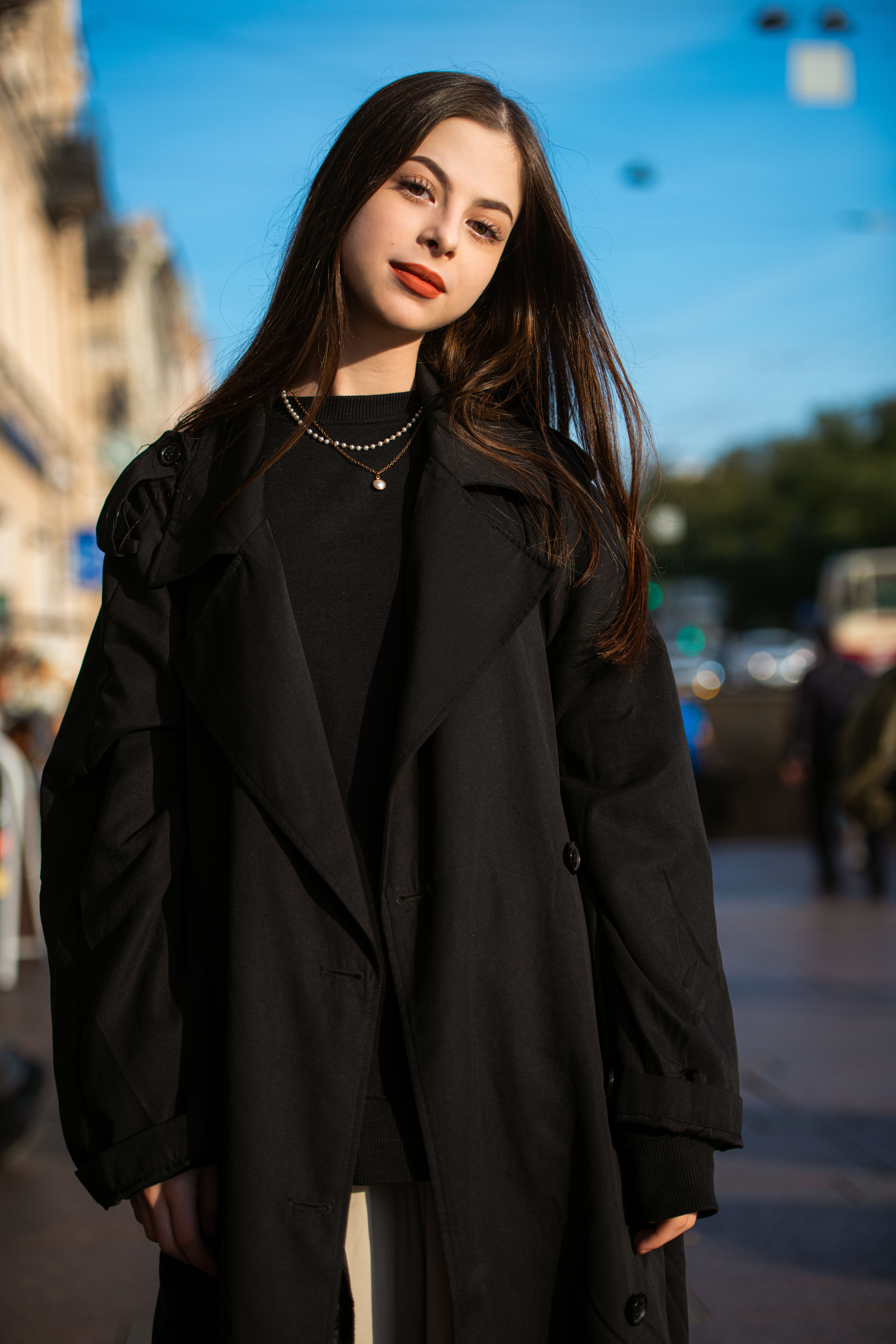 Young woman in a black coat poses stylishly on a sunlit city street.