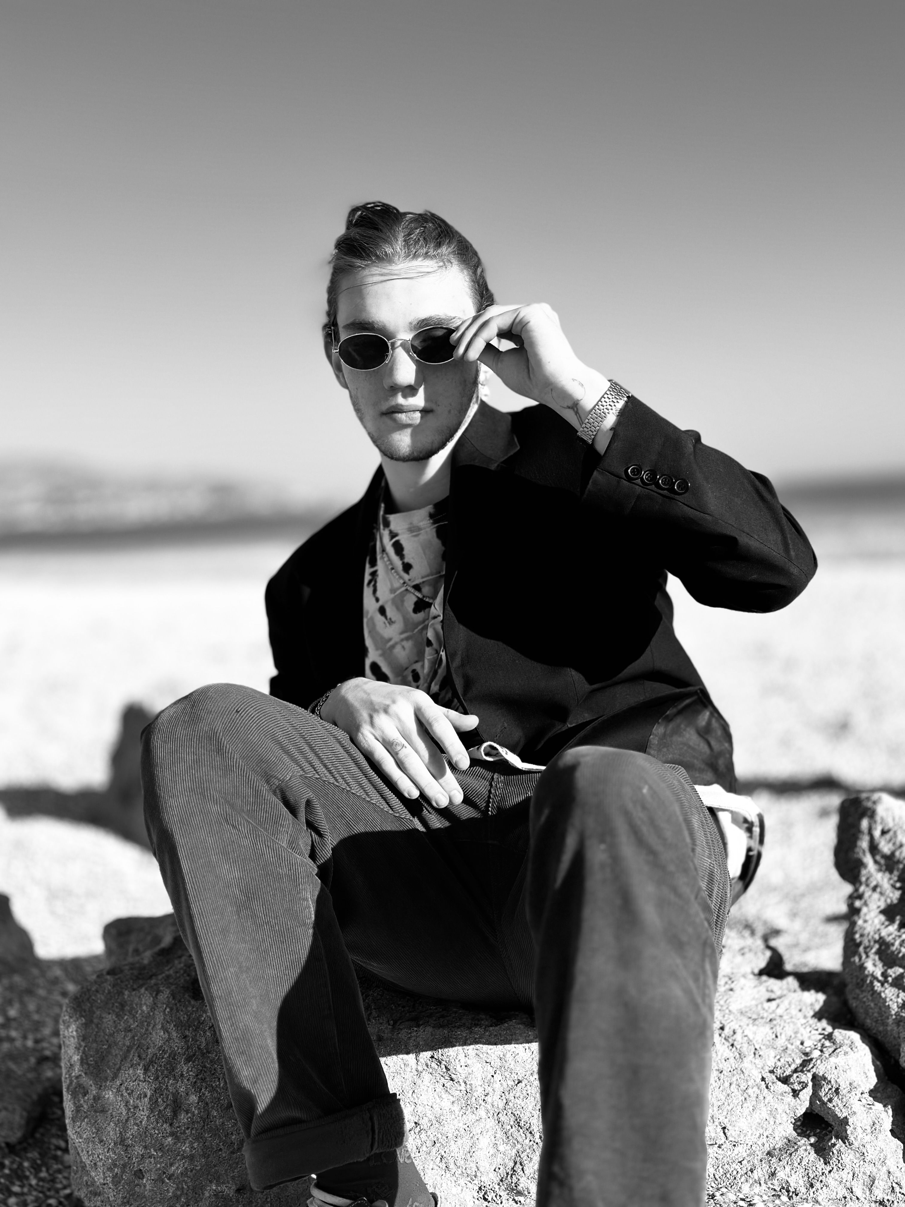 Stylish man in sunglasses posing confidently on rocks with a beach backdrop.