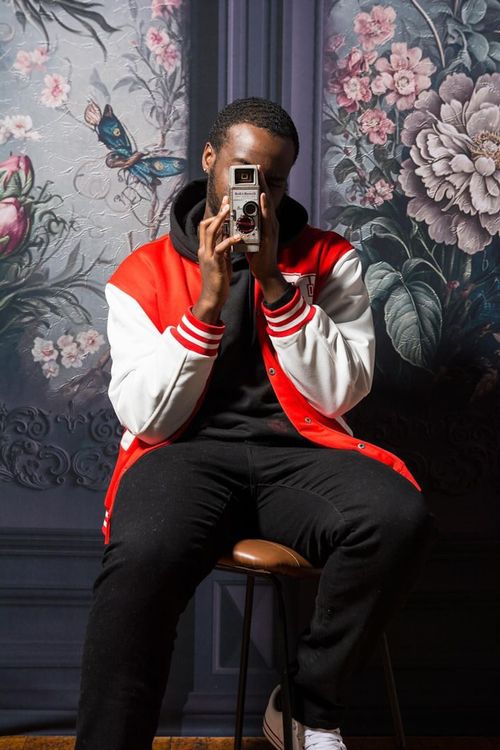 Free Fashionable man in a vintage jacket using a retro camera, seated against ornate floral wallpaper. Stock Photo