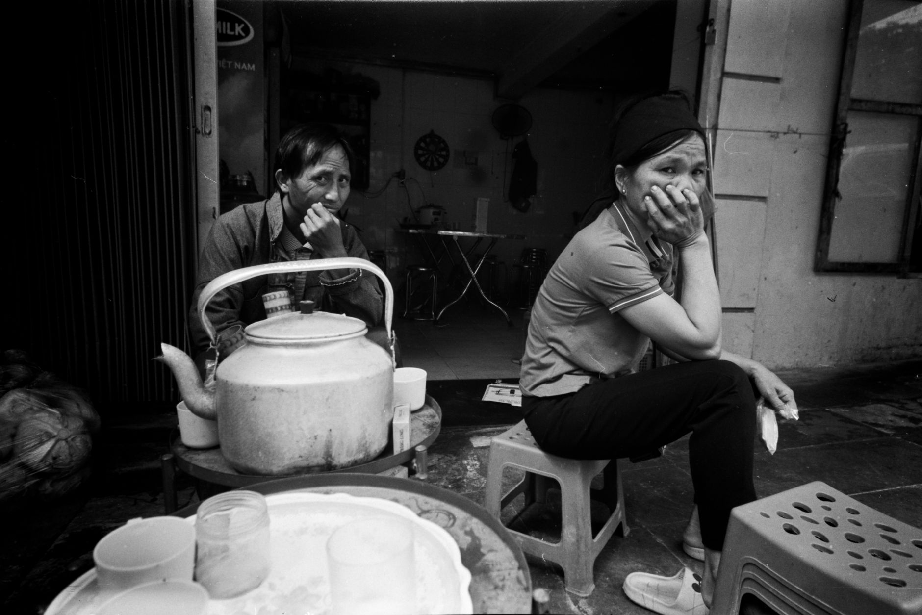 Two people sitting outside a shop with a kettle and cups, captured in black and white.
